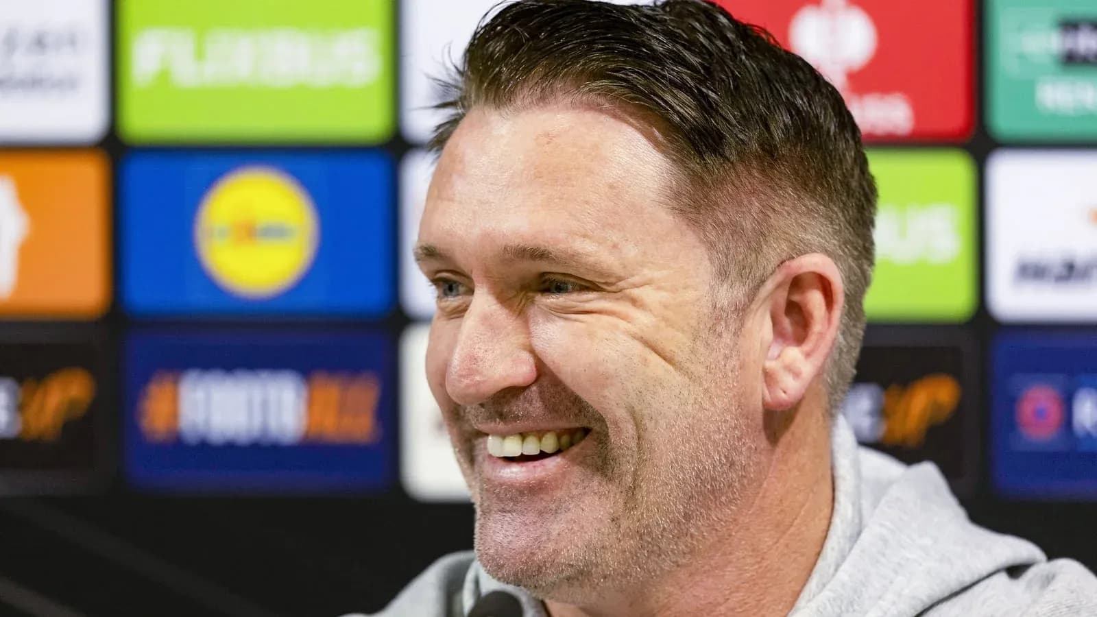 Manager smiling at press conference with logos in background