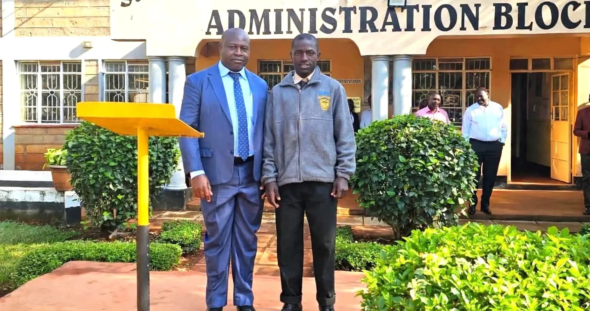Two individuals posing in front of school administration block Two individuals posing in front of school administration block